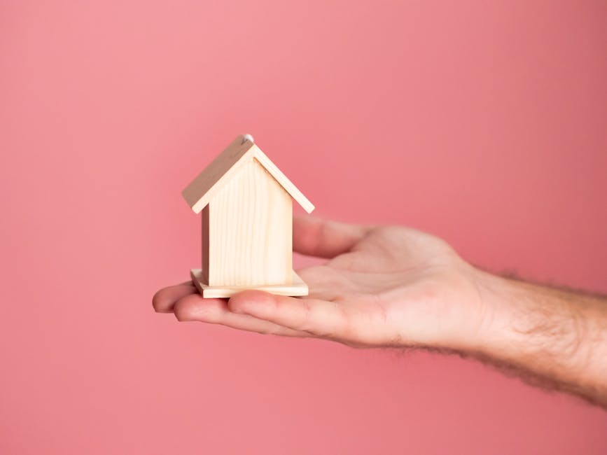 Hand holding a small wooden house model against a soft pink background, symbolizing real estate and property investment